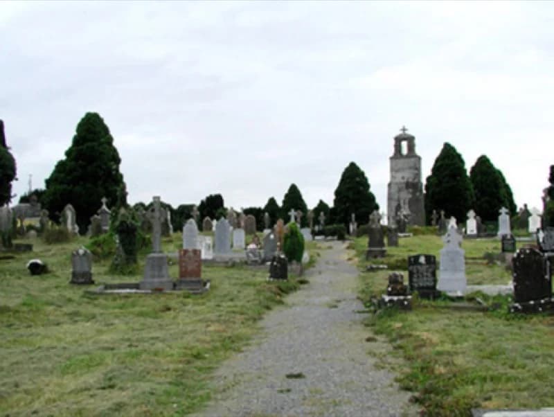 A gravel path through a graveyard of upright headstones and dark evergreens, with a pale mausoleum topped by a cross.