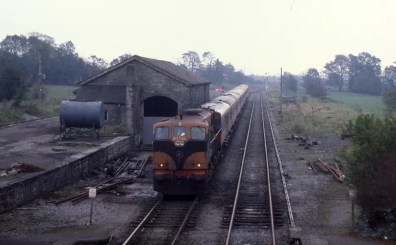 An orange and black locomotive with a train of coaches standing beside a stone goods shed at Castlerea in flat autumn light, sleepers stacked along the track.