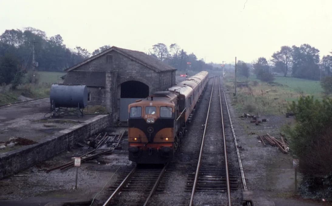 An orange and black locomotive with a train of coaches standing beside a stone goods shed at Castlerea in flat autumn light, sleepers stacked along the track.
