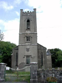 A rendered church with a battlemented three-stage tower, round-headed windows, and iron railings on stone gate piers along the road.